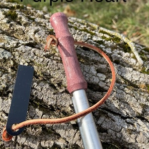 May include: A fire starter with a purple wooden handle and a silver metal body. The text "Purple Heart" is above the fire starter. A black striker and a brown leather cord are attached.