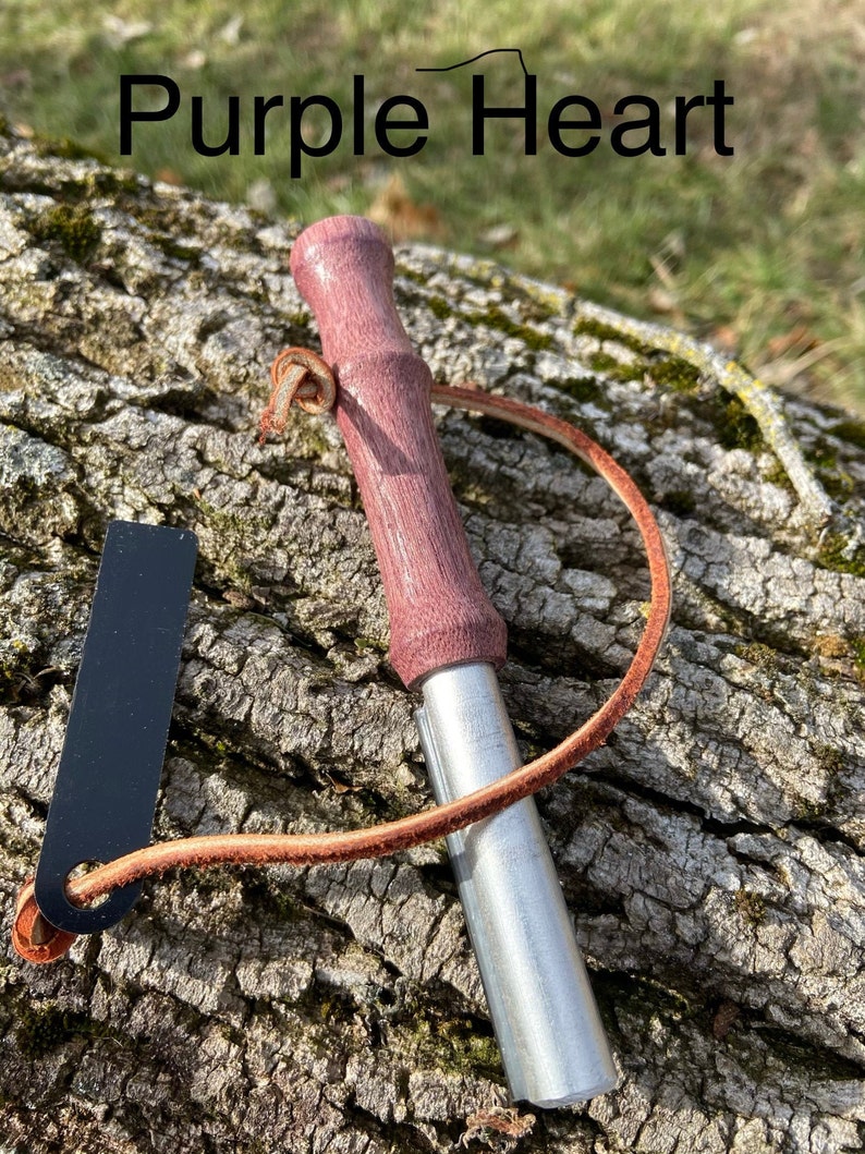 May include: A fire starter with a purple wooden handle and a silver metal body. The text "Purple Heart" is above the fire starter. A black striker and a brown leather cord are attached.