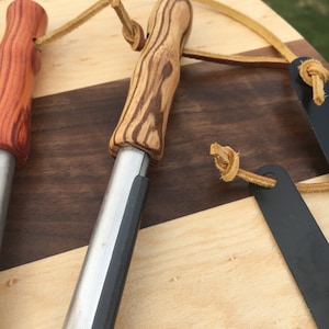 May include: Close-up of three fire starters with wooden handles and metal strikers. The handles are made of different types of wood, including a reddish-brown and a light-colored wood with dark grain. Each has a leather lanyard attached.