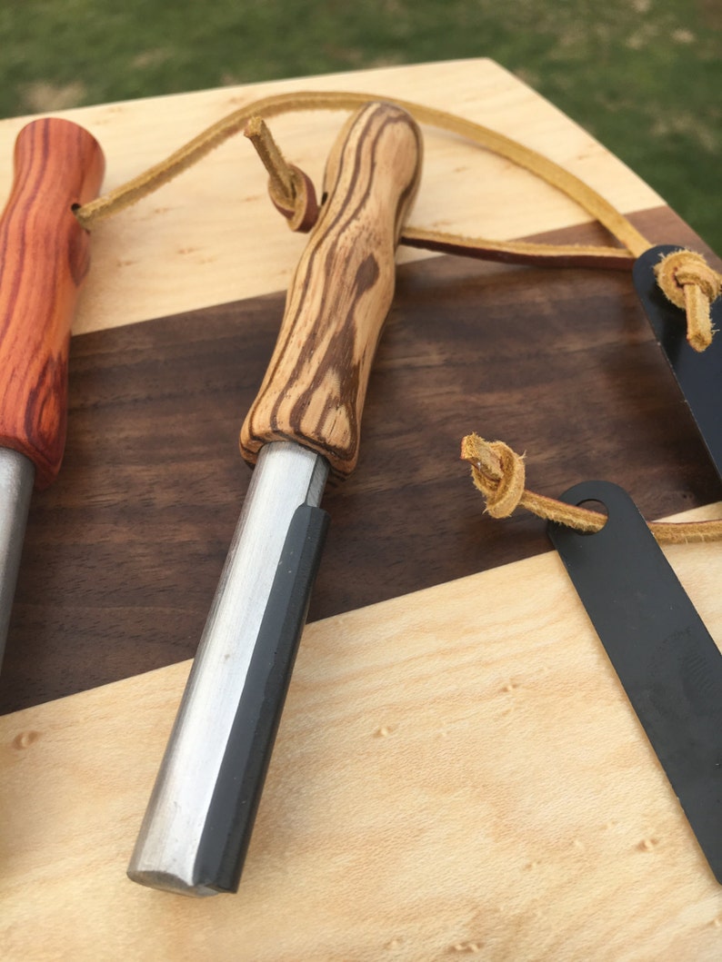 May include: Close-up of three fire starters with wooden handles and metal strikers. The handles are made of different types of wood, including a reddish-brown and a light-colored wood with dark grain. Each has a leather lanyard attached.