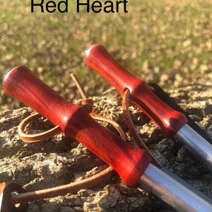 May include: Two red-handled tools with silver shafts, possibly for gardening or crafting, rest on a textured wooden surface. The handles are a deep red, and the text "Red Heart" is visible.