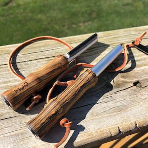 May include: Two fire starters with wooden handles and silver metal strikers. The handles have a dark wood grain pattern and are attached to leather lanyards. One handle is engraved with the initials "MJG". The fire starters are resting on a wooden surface.