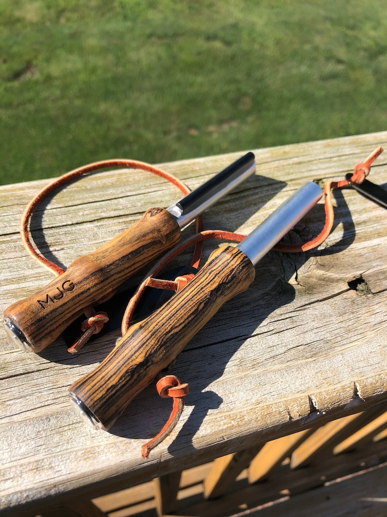 May include: Two fire starters with wooden handles and silver metal strikers. The handles have a dark wood grain pattern and are attached to leather lanyards. One handle is engraved with the initials "MJG". The fire starters are resting on a wooden surface.