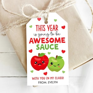 May include: A gift tag with the text "THIS YEAR is going to be AWESOME SAUCE" in red and green. The tag features two cartoon apples, one red and one green, and small red and green hearts. The tag is tied to a wrapped gift with twine.