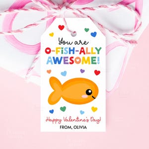以下が含まれることがあります： 「You are O-FISH-ALLY AWESOME!」の文字と漫画の金魚が描かれた白いギフトタグ。カラフルなハートと「Happy Valentine's Day!」と「FROM, OLIVIA」の文字で飾られています。