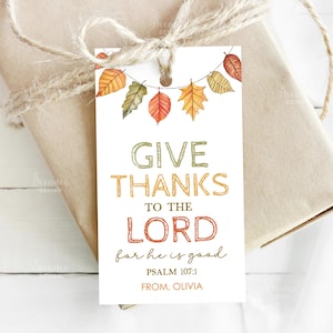 以下が含まれることがあります： 緑、オレンジ、赤で「GIVE THANKS TO THE LORD」と書かれた白いギフトタグ。秋の葉のガーランドと「PSALM 107:1 FROM, OLIVIA」の文字が特徴です。タグは紐で包まれたギフトに結ばれています。