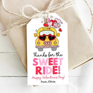 May include: A gift tag with a yellow school bus wearing heart-shaped sunglasses, and hearts floating above. The tag reads "Thanks for the SWEET RIDE! Happy Valentine's Day!" and is tied to a wrapped gift with twine.