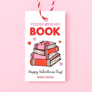 Op de afbeelding: Een wit cadeaulabel met een rood-wit touwtje. Het label bevat de tekst "YOU'RE #1 IN MY BOOK" in rood en roze, met een stapel boeken en hartjes. De onderkant van het label zegt "Happy Valentine's Day!" en "XOXO, OLIVIA".
