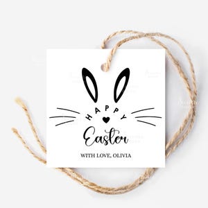 Puede incluir: Una etiqueta de regalo cuadrada blanca con un diseño de cara de conejo negro y las palabras "Happy Easter" y "With Love, Olivia". La etiqueta está atada con hilo natural.