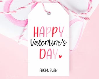 Puede incluir: Una etiqueta de regalo blanca con el texto "HAPPY Valentine's DAY" en rojo y rosa, con un pequeño corazón rojo. La etiqueta está atada con hilo rosa y blanco, lo que sugiere un tema de San Valentín.