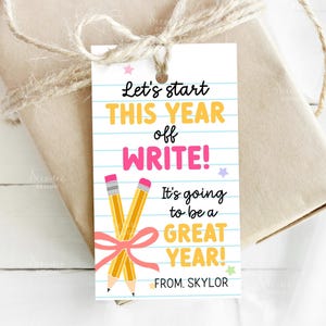 May include: A gift tag with the text "Let's start THIS YEAR off WRITE! It's going to be a GREAT YEAR! FROM: SKYLOR." The tag features two yellow pencils tied with a pink ribbon, set against a lined paper background.