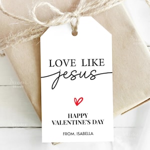 May include: A white gift tag with the text "LOVE LIKE jesus" in black, with the word "jesus" in cursive. Below is a red heart and the words "HAPPY VALENTINE'S DAY" and "FROM. ISABELLA". The tag is tied to a wrapped gift with twine.