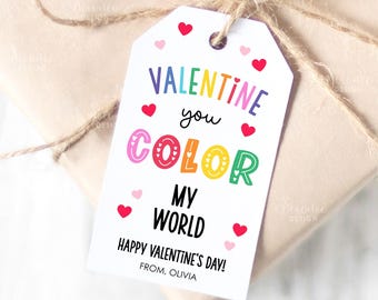 Puede incluir: Una etiqueta de regalo blanca de San Valentín con el texto "VALENTINE you COLOR MY WORLD" en letras coloridas y pequeños corazones rojos. La etiqueta está atada a un regalo envuelto con hilo.