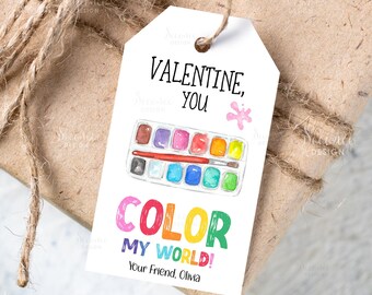 May include: A white gift tag with the text "VALENTINE, YOU" and "COLOR MY WORLD!" in colorful letters. A watercolor paint palette and brush are illustrated on the tag. The tag is tied to a wrapped gift with twine.