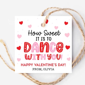 以下が含まれることがあります： 「How Sweet It Is To Dance With You!」というメッセージが赤とピンクで書かれたバレンタインデーのギフトタグ。赤いハートとピンクのハート、紐が付いた白いタグです。