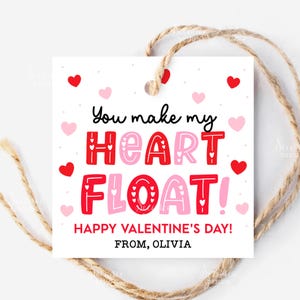 以下が含まれることがあります： 赤とピンクの「You make my HEART FLOAT!」の文字が入った、ハートの装飾が施された白い正方形のギフトタグ。タグには「HAPPY VALENTINE'S DAY! FROM, OLIVIA」と書かれており、バレンタインデーに最適な麻ひもで結ばれています。