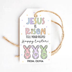 以下が含まれることがあります： 「JESUS is RISEN TELL YOUR PEEPS! Happy Easter!」の文字と3匹のウサギのイラストが描かれた白いイースターギフトタグ。タグには吊り下げ用のジュート紐が付いており、下部には「OLIVIA」の名前が記載されています。