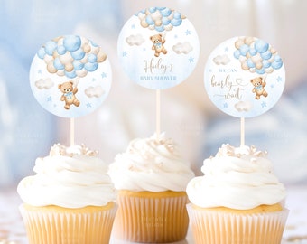 May include: Three cupcakes with white frosting topped with blue and beige balloon cupcake toppers featuring a teddy bear holding a balloon. The toppers say "Hailey's Baby Shower" and "We can bearly wait".