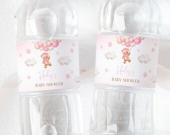 May include: Two clear plastic water bottles with pink and gold labels. The labels feature a teddy bear holding pink and gold balloons, clouds, and snowflakes. The text on the labels reads "Hailey's Baby Shower".