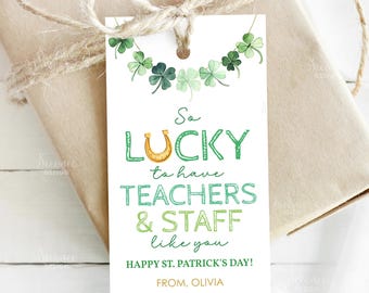 May include: A white St. Patrick's Day gift tag with green text that reads "So Lucky to have TEACHERS & STAFF like you." The tag has a horseshoe and shamrock garland design. The tag is tied to a wrapped gift with twine.