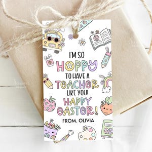 以下が含まれることがあります： カラフルな文字で「I'M SO HOPPY to have a TEACHER LIKE YOU! HAPPY EASTER! FROM. OLIVIA」と書かれたギフトタグ。スクールバス、花、虹、その他の学校をテーマにしたグラフィックのイラストが特徴です。