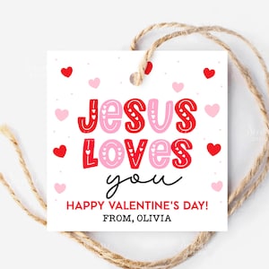 May include: A white Valentine's Day gift tag with the message "JESUS LOVES you" in red and pink lettering, surrounded by red and pink hearts. The tag also includes the text "HAPPY VALENTINE'S DAY! FROM, OLIVIA" and is attached to a twine string.