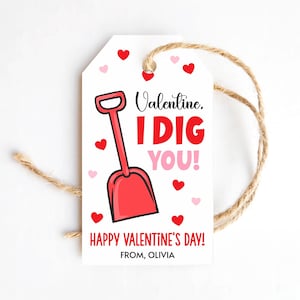 May include: A white Valentine's Day gift tag with a red shovel illustration and the text "Valentine. I DIG YOU!" in red and pink. The tag also includes the text "HAPPY VALENTINE'S DAY!" and "FROM, OLIVIA", with red and pink heart accents and a twine string.