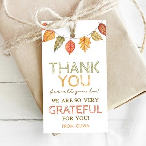 May include: A white gift tag with a string of colorful autumn leaves. The tag reads "THANK YOU for all you do! WE ARE SO VERY GRATEFUL FOR YOU! FROM, OLIVIA" in various colors and fonts. The tag is tied to a wrapped gift with twine.