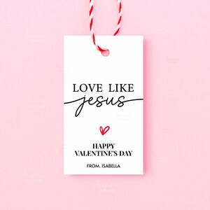 Pode incluir: Uma etiqueta de presente branca com o texto "LOVE LIKE jesus" em escrita preta, um pequeno cora&ccedil;&atilde;o vermelho e "HAPPY VALENTINE'S DAY" abaixo. A etiqueta est&aacute; presa a uma corda vermelha e branca, em um fundo rosa.