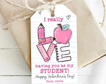 May include: A gift tag with a pencil, ruler, apple, and notebook design spelling "LOVE". The tag reads "I really having you as my STUDENT! Happy Valentine's Day! from. olivia". The tag is tied to a wrapped gift with twine.