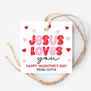 May include: A white Valentine's Day gift tag with the message "Jesus Loves You" in red and pink lettering, surrounded by hearts. The tag includes the text "Happy Valentine's Day! From, Olivia" and is tied with twine.