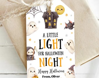 May include: A Halloween gift tag with a spooky theme. The tag features a haunted house, ghosts, a bat, and the text "A Little Light for Halloween Night." The tag is tied to a wrapped gift with twine.