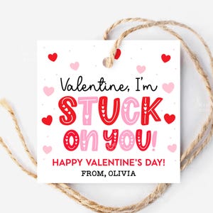 May include: A square white Valentine's Day gift tag with the text "Valentine, I'm STUCK on you!" in red and pink. The tag is decorated with red and pink hearts and includes the message "HAPPY VALENTINE'S DAY! FROM, OLIVIA". A natural twine string is attached.