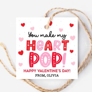 May include: A white square gift tag with the text "You make my HEART POP!" in red and pink. The tag includes the message "HAPPY VALENTINE'S DAY! FROM, OLIVIA" and is strung with twine, with red and pink hearts scattered around.