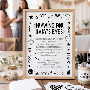 May include: A framed sign with the text "DRAWING FOR BABY'S EYES" and instructions for creating black and white art for babies. Includes a stack of white cards, black markers, and sample designs. A baby shower theme.