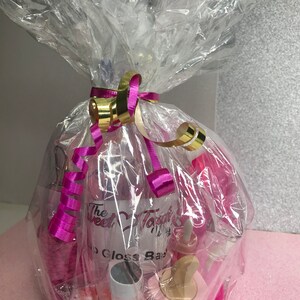 May include: A gift basket wrapped in clear cellophane, tied with a gold and pink ribbon. The basket contains various pink lip gloss products, including bottles and containers. The text "The Sweet Topic" is visible on a bottle.