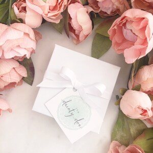 May include: A white gift box tied with a white ribbon and a tag that reads "the dainty doe." The box is surrounded by pink flowers with green leaves, creating a soft, feminine aesthetic. The tag has a light green circular design.