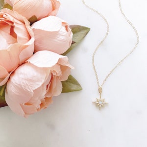 May include: A gold necklace featuring a starburst pendant with a central round stone and small clear stones set along the points. The delicate gold chain is attached to the pendant, which is positioned next to a cluster of light pink peony flowers.