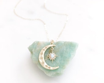 May include: A gold-toned necklace featuring a crescent moon pendant engraved with small star motifs. A dangling starburst charm with a central oval opal-like stone and tiny clear accents hangs from the center of the crescent moon pendant.