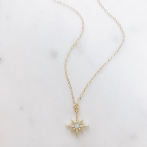 May include: A gold-toned necklace featuring a delicate chain and a starburst-shaped pendant. The pendant is accented with small clear stones and a central iridescent round stone, creating a radiant, multi-pointed star design with a polished finish.