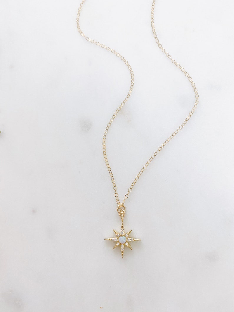 May include: A gold-toned necklace featuring a delicate chain and a starburst-shaped pendant. The pendant is accented with small clear stones and a central iridescent round stone, creating a radiant, multi-pointed star design with a polished finish.