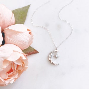 K&ouml;nnte beinhalten: Eine silberne Halskette mit einem Halbmond-Anh&auml;nger und einem Stern-Anh&auml;nger mit einem Opal-Stein. Die Halskette liegt auf einem wei&szlig;en Hintergrund mit rosa Blumen.