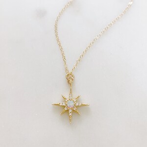 May include: A gold-toned necklace featuring a delicate chain and a starburst-shaped pendant. The pendant is centered with a small, iridescent round opal stone and is accented with tiny, clear pav&eacute;-set crystals along each of the eight radiating points.