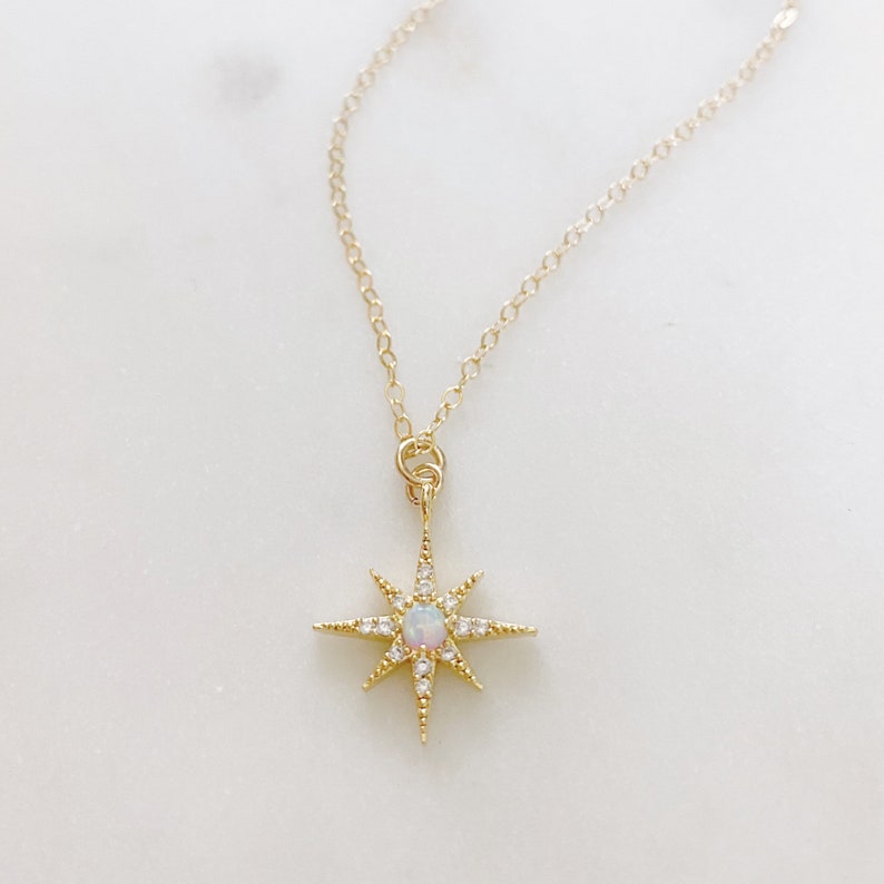 May include: A gold-toned necklace featuring a delicate chain and a starburst-shaped pendant. The pendant is centered with a small, iridescent round opal stone and is accented with tiny, clear pav&eacute;-set crystals along each of the eight radiating points.