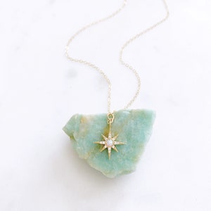 May include: A gold-toned necklace featuring a starburst pendant with a central iridescent stone and small clear accents. The pendant is suspended from a delicate gold-toned chain and rests against a rough, light green mineral stone piece.