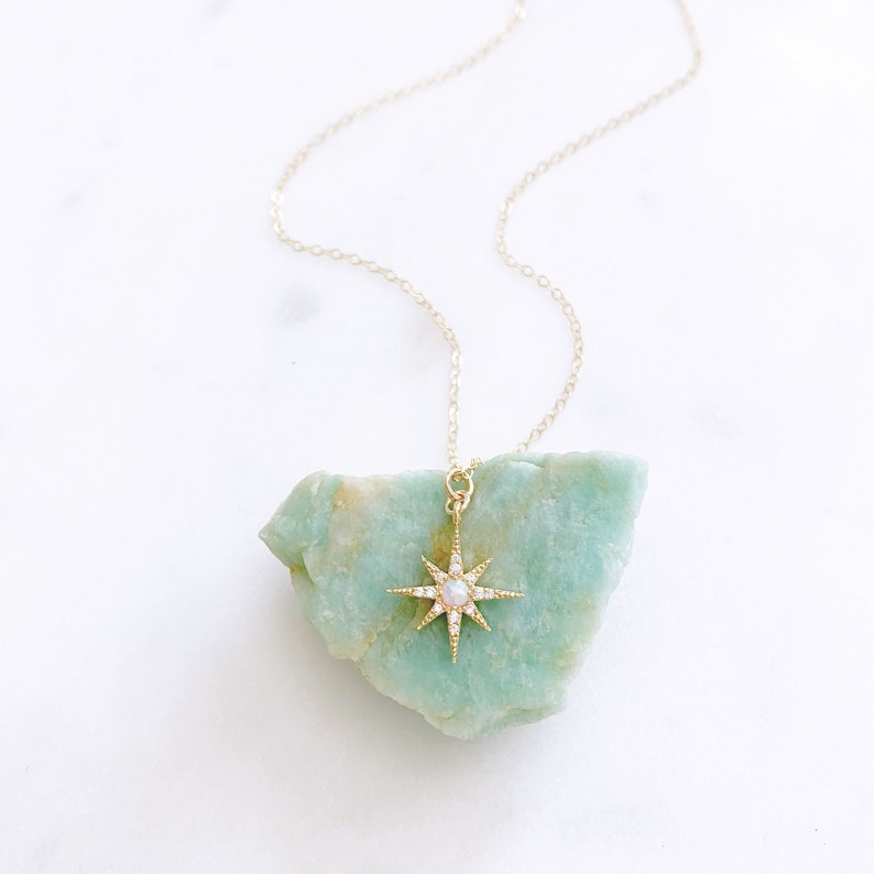 May include: A gold-toned necklace featuring a starburst pendant with a central iridescent stone and small clear accents. The pendant is suspended from a delicate gold-toned chain and rests against a rough, light green mineral stone piece.