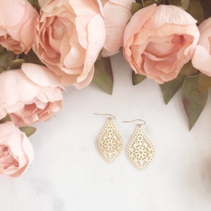 May include: A pair of gold-colored, filigree teardrop earrings with a floral design. The earrings are displayed on a white surface, surrounded by soft pink flowers.