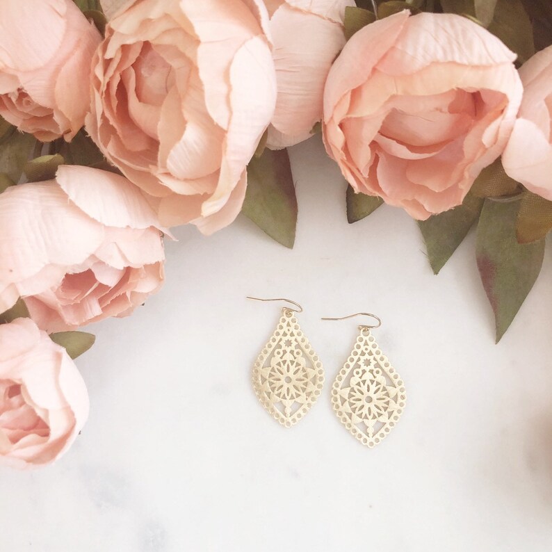 May include: A pair of gold-colored, filigree teardrop earrings with a floral design. The earrings are displayed on a white surface, surrounded by soft pink flowers.