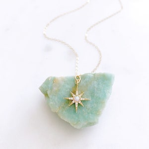 May include: A gold necklace featuring a starburst pendant with a central round opal stone and small clear pav&eacute; crystals along the rays. The pendant is suspended from a delicate gold chain, showcasing a bright, reflective metallic finish.