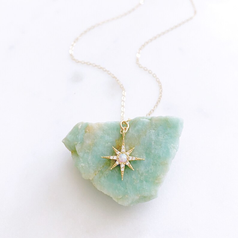 May include: A gold necklace featuring a starburst pendant with a central round opal stone and small clear pav&eacute; crystals along the rays. The pendant is suspended from a delicate gold chain, showcasing a bright, reflective metallic finish.